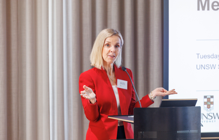 A woman in a red jacket speaks next to a lectern